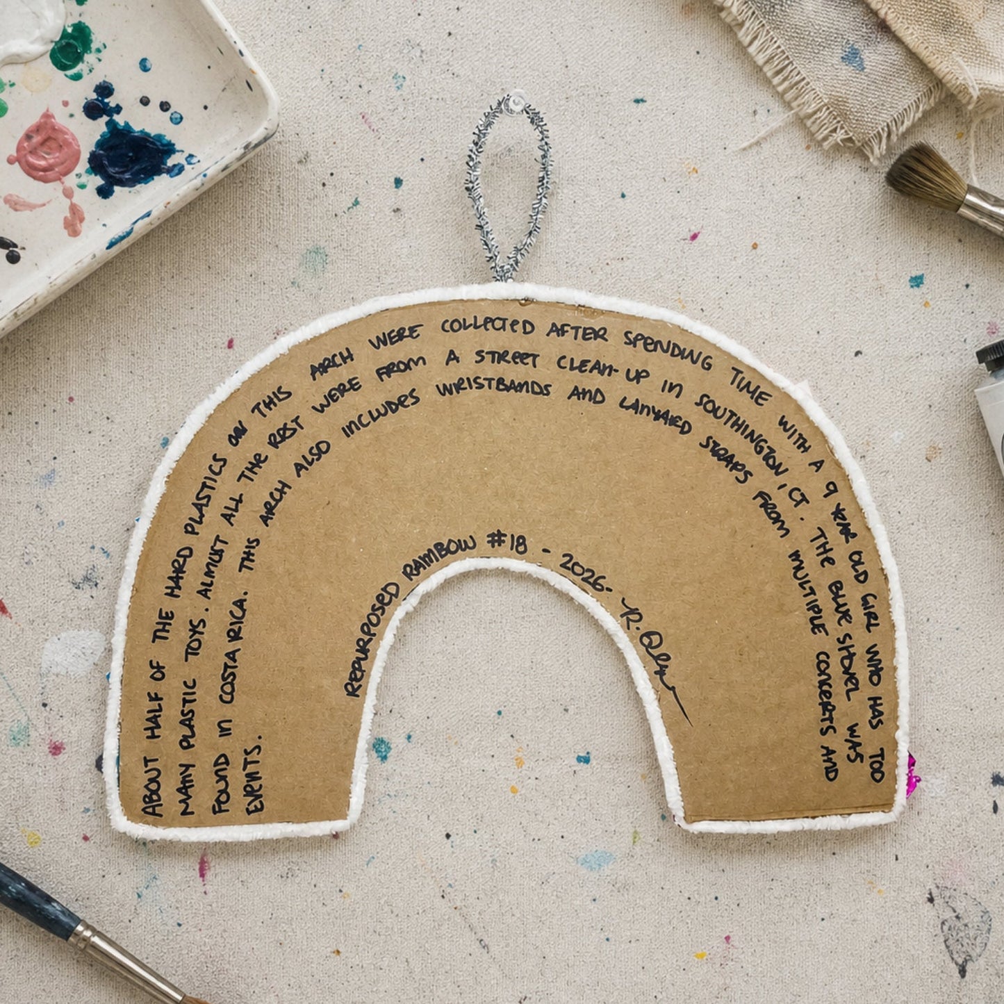 Handmade rainbow-shaped craft item with text on a textured surface with art supplies.